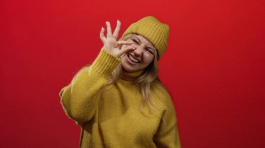 Sarı kazaklı ve bereli bir kadın kırmızı arka planda gülümsüyor. Elleriyle iyi bir jest yapıyor, sıcaklığını ve pozitifliğini gösteriyor..