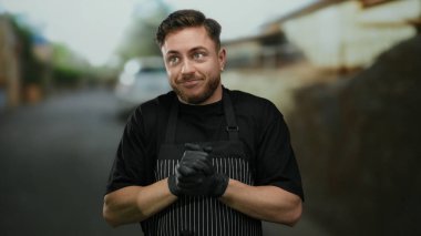 Sakallı, önlüklü, eldivenli, beyaz bir genç, açık bir caddede duruyor, garson kılığında..