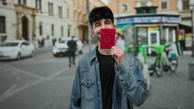 Kalabalık bir şehir caddesinde İtalyan pasaportu taşıyan mutlu bir adam, açık şehir ortamında seyahat etmeye hazır ve neşelidir, parlak bir gülümseme ve günlük kıyafetlerle.