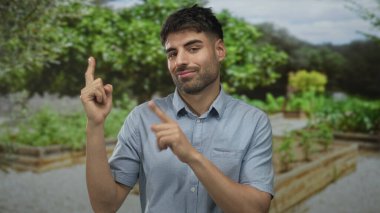 Mavi gömlekli genç İspanyol adam, bulutlu gökyüzünün altındaki yeşil parktaki yükseltilmiş bahçe yatağının yakınındaki ağaca işaret ediyor..