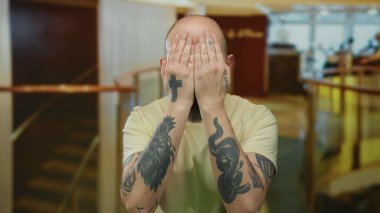 Sakallı adam, bir otel lobisinde şakacı bir şekilde yüzünü örten, karizma sergileyen ve kapalı mekanda çekici bir kişiliğe sahip..