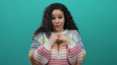 Renkli süveter giyen bir kadın yeşil arka plana karşı heyecan ve şaşkınlık dolu kıvırcık saçlar ve neşeli bir gülümseme sergiliyor..