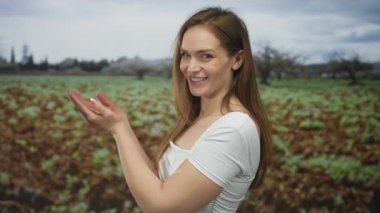 Genç beyaz kadın bulutlu gökyüzü altında tomurcuklanan bitkilerin arasında yeşil alanda ellerini ileri uzatıyor; dinginlik.