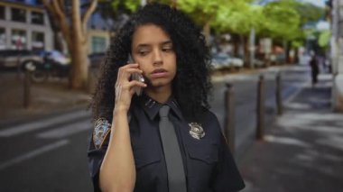 Üniformalı kadın polis telefonda konuşuyor. Sokakta, dışarıda ağaçlarla kurulmuş ve arka planda bulanıklık var..