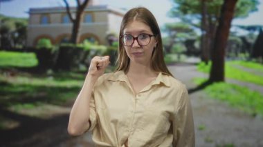 Ağaçlar ve çitlerle çerçevelenmiş avluda, gözlüklü genç sarışın kadın göğsüne yakın yumruğunu sıkıyor;.