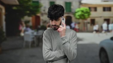 Genç bir adam, düşünceli bir ifadeyle, gri bir kazak giymiş, canlı, hareketli bir şehir manzarasının arka planına karşı akıllı bir telefonla konuşuyor..