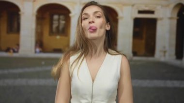 Güneşli eski kasaba avlusunda taş kemerlerle gezen bir kadın kameraya dil uzatıyor..