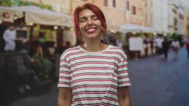 Kızıl saçlı ve çizgili tişörtlü bir kadın. Elleri önde, bilezik ve küpelerle gülümsüyor. Teras restoranının önünde duruyor..