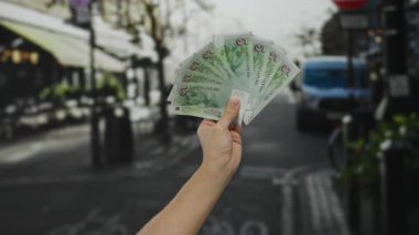 El ele tutuşan Singapur Dolarları, açık şehir arka planına sahip. Park etmiş araçlar ve bulanık kafeler ile hareketli sokak sahnelerini gösteriyor.