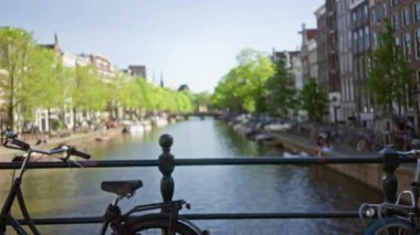 Güneşli bir günde yemyeşil ağaçların çerçevelediği köprünün üstünde bisikletleri ve tarihi binaları olan Amsterdam Kanalı 'nın bulanık görüntüsü..