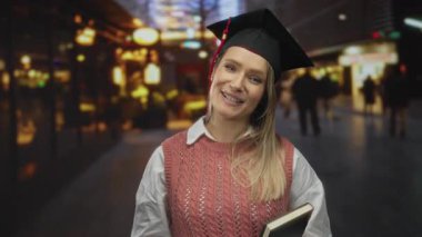 Mezuniyet şapkası içinde gülümseyen bir kadın, şehir sokaklarında kitap tutuyor, şehir yaşamında neşe ve başarıyı somutlaştırıyor, açık havada eğitimi kutluyor..