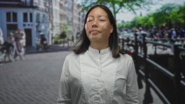 Gözleri kapalı bir kadın yoğun bir şehir caddesinde duran çilli yüzünü gösteriyor arkasında kanallar var; sakin yansıma.