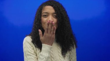 Parlak mavi stüdyo ortamında gülümserken el ele öpüşen Afrikalı Amerikalı bir kadın..