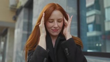 Şehir merkezindeki kadın, şehir merkezinin arka planında siyah ceket giyen ve rahatsızlığı veya baş ağrısını gösteren kapalı gözlerle başını tutuyor.