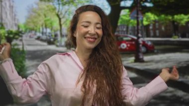 Elleri çerçeveli gülümseyen, pembe tişörtlü ve uzun kıvırcık saçlı bir kadın, park halindeki arabalarla yapraklı bir şehir caddesinde poz veriyor;.