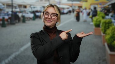 Yüzü bulanık şehir arka planına sahip, gülümseyen ve parmakla işaret eden bir kadın, açık bir şehir manzarası yakalıyor, genç sarışın kadın bir şeyi vurguluyor..