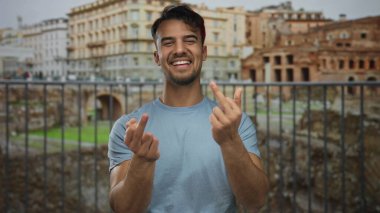 Hispanik bir adam orta parmaklarıyla hareketler yaparken açık bir şehirde Roma harabelerinin önünde gülüyor mizah ve saygısızlıklar sergiliyor..