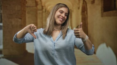 Sarışın genç kadın, tarihi ön cephede başparmağıyla işaret ediyor ve baş parmağıyla yere eğiyor; belirsizlik.