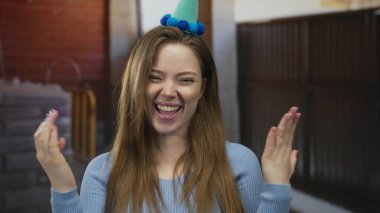 Parti şapkası takan kadın, bina içinde dişlerini gösteren el kaldırıyor ve gülümsüyor; doğum günü neşesi.