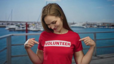 Kadın deniz kıyısındaki açık kırmızı gönüllü gömleğini işaret ediyor; gönüllü toplum hizmeti gururu.