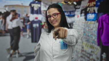 Beyaz üniformalı bir kadın, elleri açık pazar ortamında alışveriş yapanlar ve renkli mallar arasında gülümseyen bir kalp oluşturur..