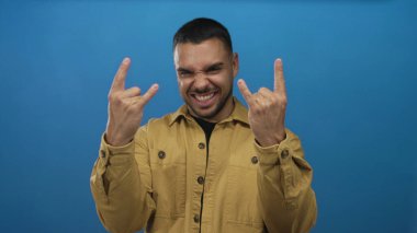 İnsan elleri stüdyoda kameraya geniş bir şekilde gülümserken mavi zemine karşı rock boynuzları hareketi oluşturur; eğlenceli müzik enerjisi titreşimi.