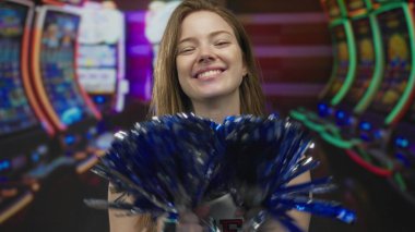 Genç bir kadın kumarhane binasında kumar makinesinin önünde tezahürat yaparken mavi ve gümüş ponponu sallıyor..