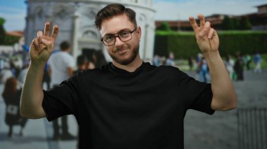 İtalya 'da sidik kulesinin önünde barış işareti yapan, arka planda kalabalık olan gözlük takan, seyahat ve turizm havası gösteren sakallı genç bir adam.