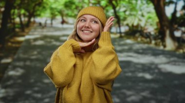 Hardal süveter ve şapkayla gülümseyen bir kadın, yemyeşil ağaçlarla dolu güneşli bir parkta mutlu poz veriyor..