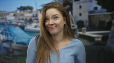 Genç bir kadın güneş ışığı binasında mavi teknelerin yanında el kaldırarak gülüyor ve jestler yapıyor..