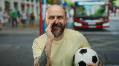 Sakallı, sakallı, elinde futbol topu olan, otobüslü ve arka planda yürüyen bir adam..