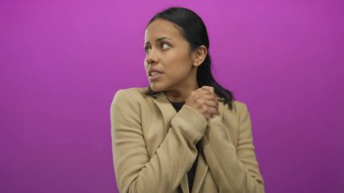 Kısa koyu renk saçlı ve bej ceketli İspanyol bir kadın soyutlanmış pembe bir arkaplan karşısında endişeli görünüyor, elleri göğsüne yapışık, anksiyete ve tefekkür taşıyor..