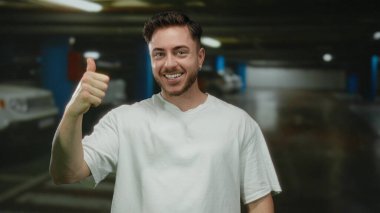 Arka planda bulanık arabalarla kapalı bir otoparkta dikilirken sakallı genç adam gülümsüyor ve başparmak kaldırıyor..