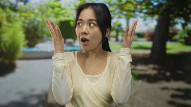 Mavi gökyüzünün altındaki yemyeşil ağaçlar ve patikalarla çevrili yeşil bir park ortamında şaşkınlığını ifade eden bir kadın..