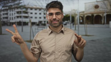 Dost canlısı gülümseyen gözlüklü genç bir adam ve iki eli sokakta barış işareti gösteren bej bir gömlek..