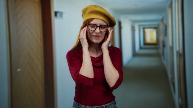 Gözlüklü ve kızıl saçlı bir kadın düşünceli bir şekilde bir otel koridorunda duruyor, kırmızı bir kazak ve sarı bereli, düşünce ve stil ifadelerini yakalıyor..