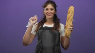 Mor stüdyoda açık plastik eldiven ve çizgili önlük giyerken elinde uzun bir baget ve işaret parmağı tutan kadın fırıncı; neşeli ferahlık.