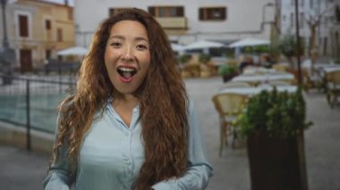 Elleri görünür şekilde gülümseyen, açık mavi gömlekli ve uzun saçlı bir kadın, açık hava masalarının yanındaki bir restoranın terasında duruyor; neşe bağlantısı.