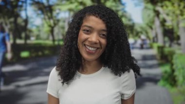 Afrika kökenli Amerikalı kadın, güneş ışığında kameraya gülümserken yeşil park yolunda elini yüzünü kapatıyor..