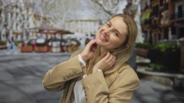 Şehir sokaklarında gülümseyen bir kadın, sarı saçlı, bej ceketli, açık renk elbiseli, neşeli ve sıradan bir şehir manzarası sergiliyor..