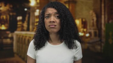 Kadın, loş aydınlatılmış kilise binasında süslü katedral sunağı önünde kaşlarını çatar; belirsizlik iç gözlem.