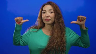 İki eliyle parmaklarını işaret eden ve mavi stüdyoda yeşil kazak ve uzun saç giyen, kendinden şüphe eden bir kadın..