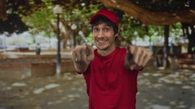 Kırmızı gömlekli ve şapkalı bir adam yeşil yaprakların arasında akıllı telefonu tutarken kamerayı işaret ediyor..