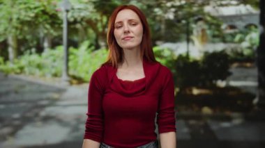 Şehir dışındaki kadın, kızıl saçlı ve çekici, günlük kırmızı bluz giyiyor, yeşil ve güneş ışığıyla çevrili, kentsel ortam, duygularını canlı bir şekilde ifade ediyor..