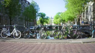 Bisikletler Amsterdam 'da bir kanal boyunca dizilmiş. Arka planda bulanık ağaçlar ve suyun kentsel zarafeti yansıtması var..