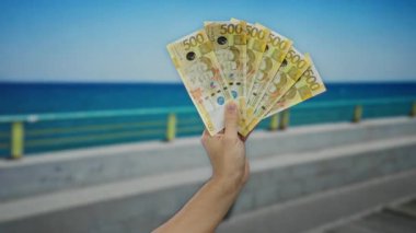 Arka planda açık mavi gökyüzü olan deniz kenarındaki gezinti alanının yanında Filipinli pesoları tutan adam plajda rahatlamış bir havayla para sergiliyor..