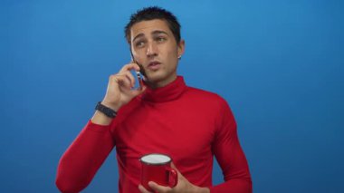 Stüdyoda kahve fincanı tutarken kırmızı süveter giyen genç İspanyol adam akıllı telefonda konuşuyor..