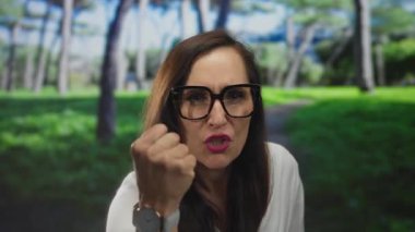 Gözlüklü bir kadın, güneşli bir günde, açık yeşil bir parkta, uzun ağaçlarla çevrili bir yerde, net bir şekilde odak ve ifadesini gösteriyor..