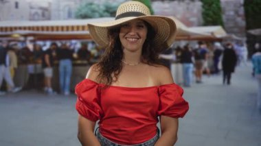 Gülümseyen bir kadın, elinde saman güneş şapkası, çıplak omuzlar görünür, kırmızı omuzlu, kalabalık bir cadde pazarında, yaz seyahati neşesi.