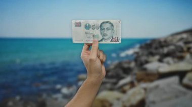 Resimli bir plaja karşı elinde Singapur Doları banknotları tutan adam huzurlu bir sahil ortamında seyahat ve finansal özgürlüğün altını çiziyor..